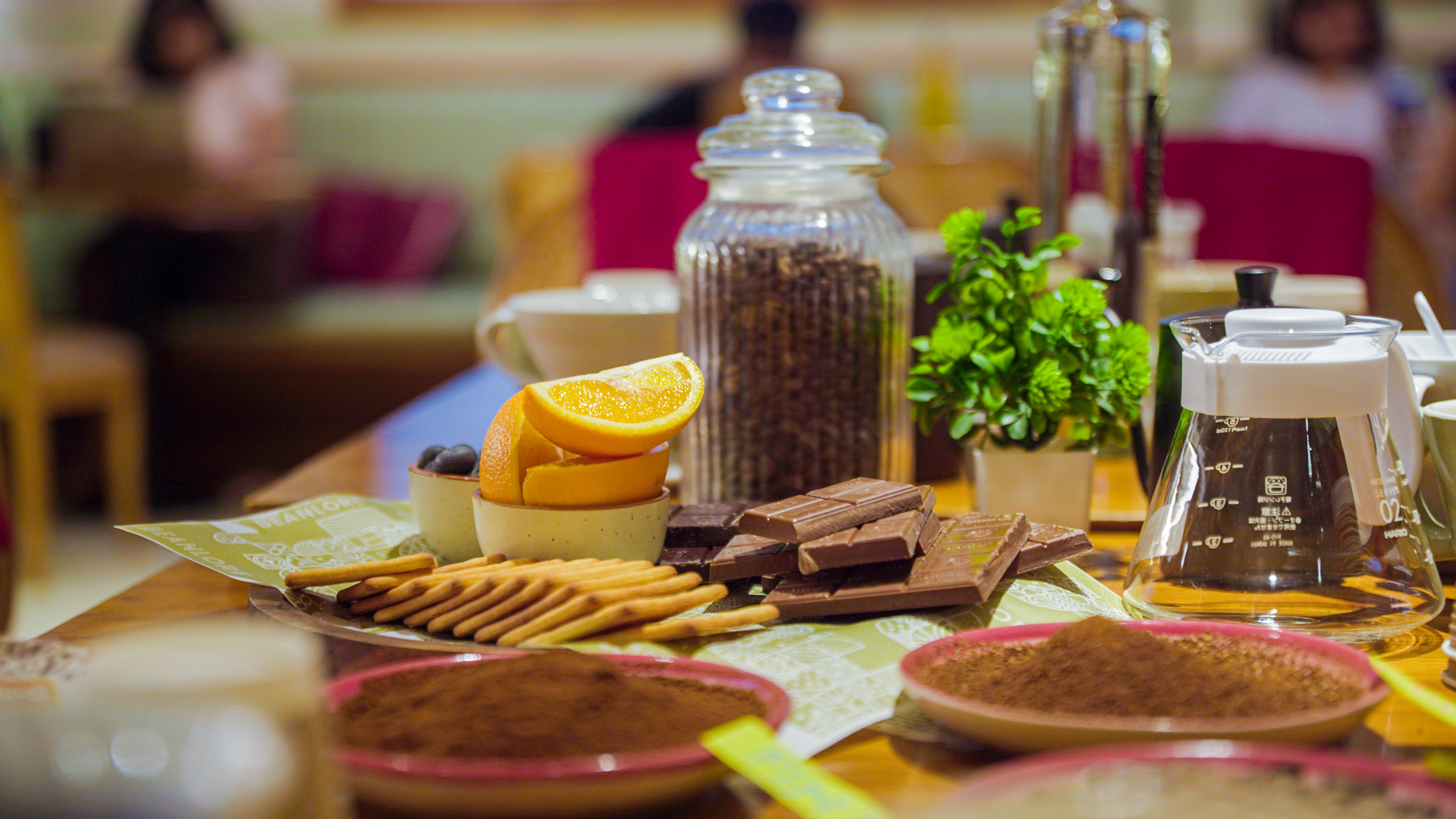 Coffee beans in a jar, chocolate bars, orange slices, biscuits and coffee-related items on a wooden table.